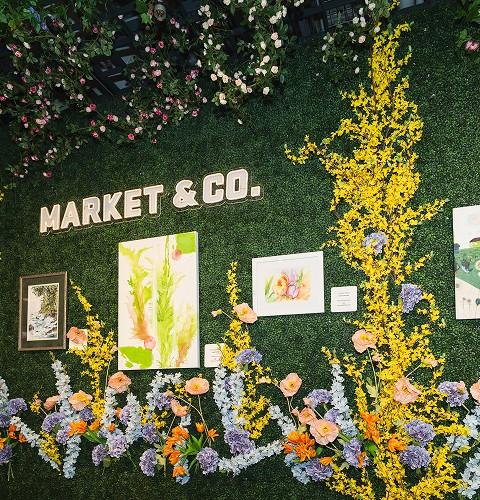 Floral Market & Co. art display on a green hedge wall with paintings