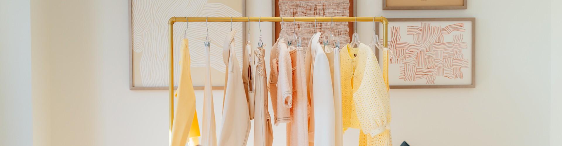 Rack of pastel yellow and cream clothing below framed art in a boutique