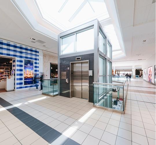 Glass elevator in a sunlit mall concourse near a blue gingham storefront
