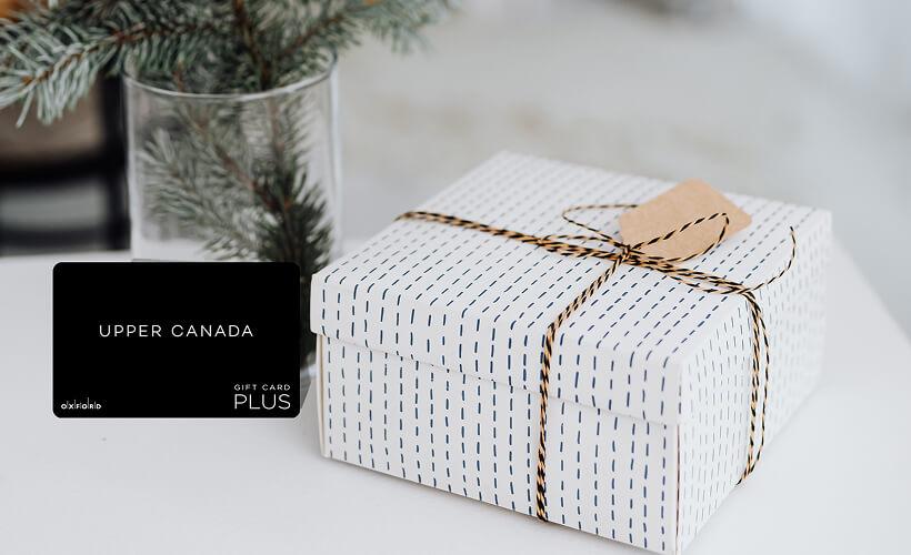 Black Upper Canada Plus gift card beside a wrapped white gift box and pine