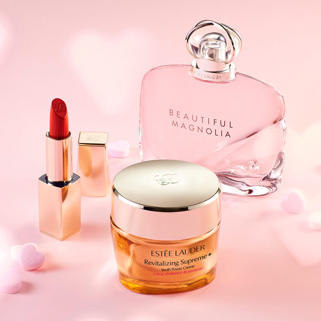 Estee Lauder lipstick, cream, and perfume are positioned on a light pink background.