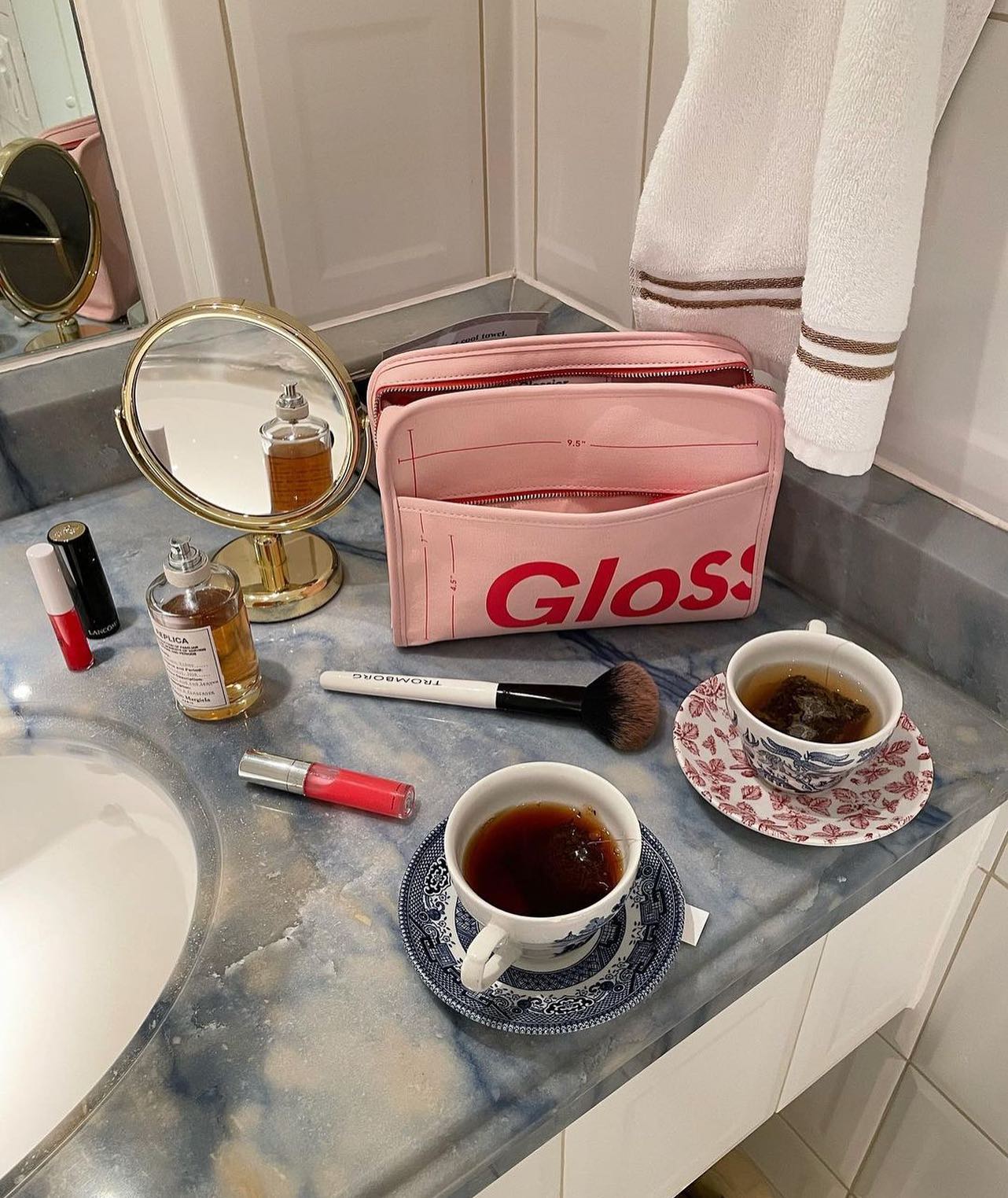 A Glossier makeup bag sits on a bathroom counter top alongside other Glossier products and two cups of tea.