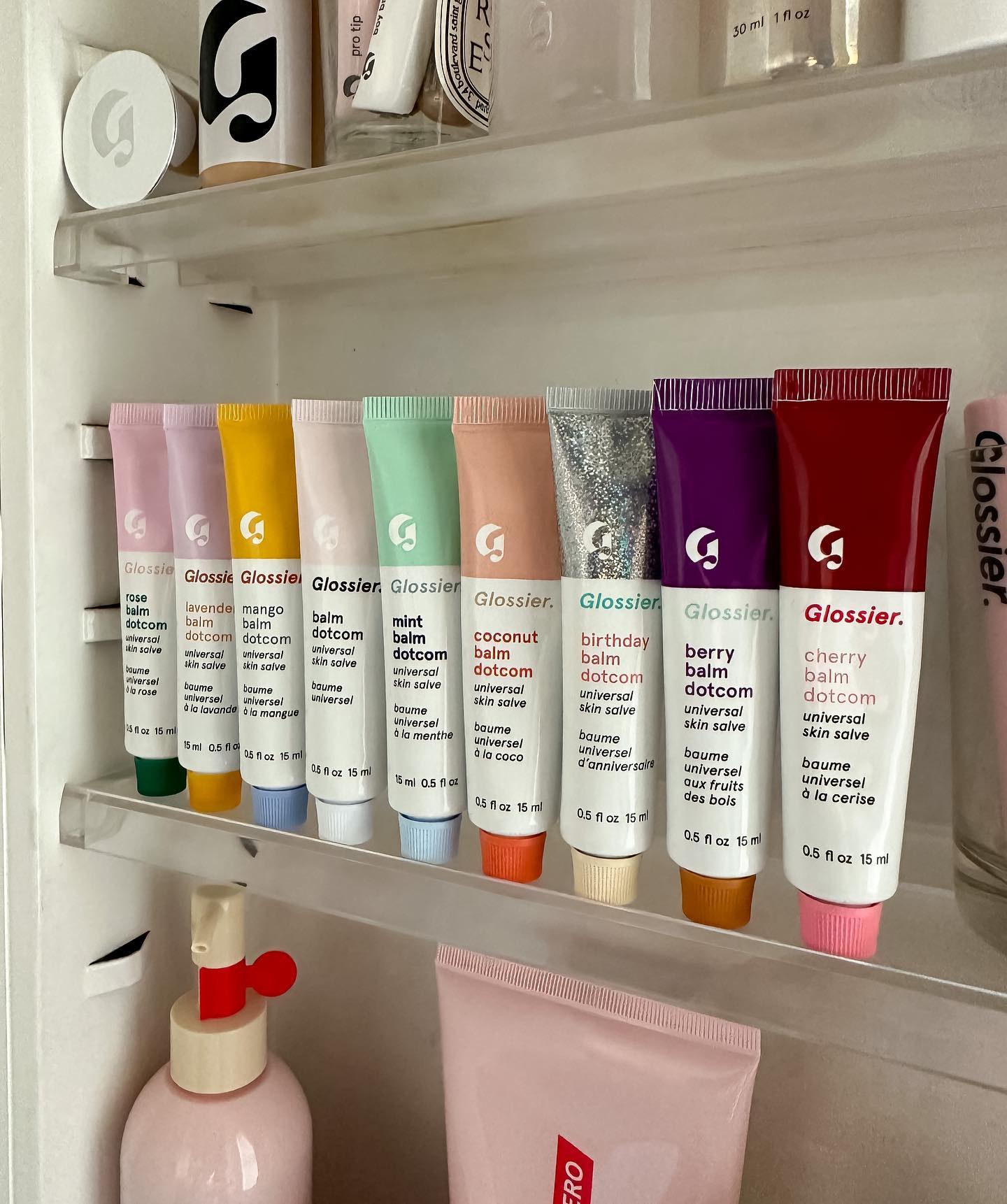 Various colours of Glossier lip gloss are set up in a line within a bathroom vanity cabinet.