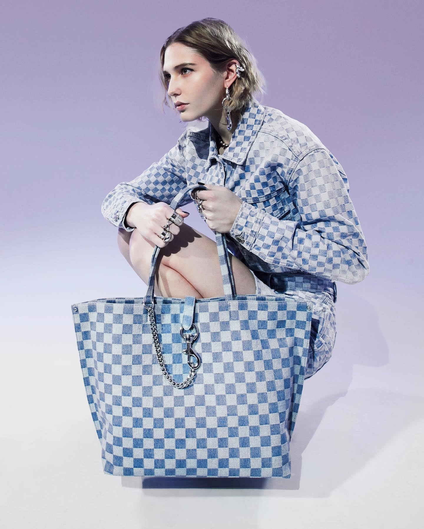 A woman bends at the knee while holding a blue checkered bag from Rebecca Minkoff. Her jacket matches the bag.