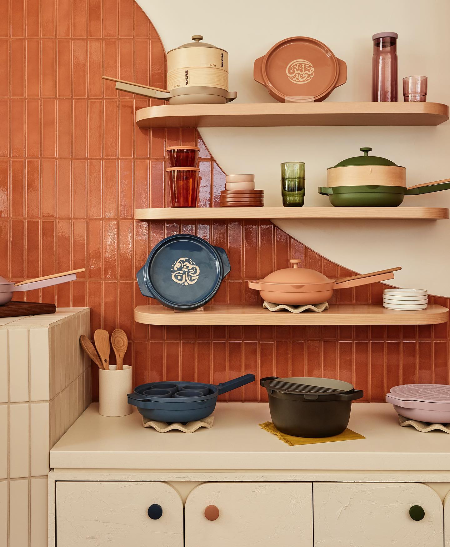 Various colours of Our Place pots and pans are displayed on a retro-inspired wall.