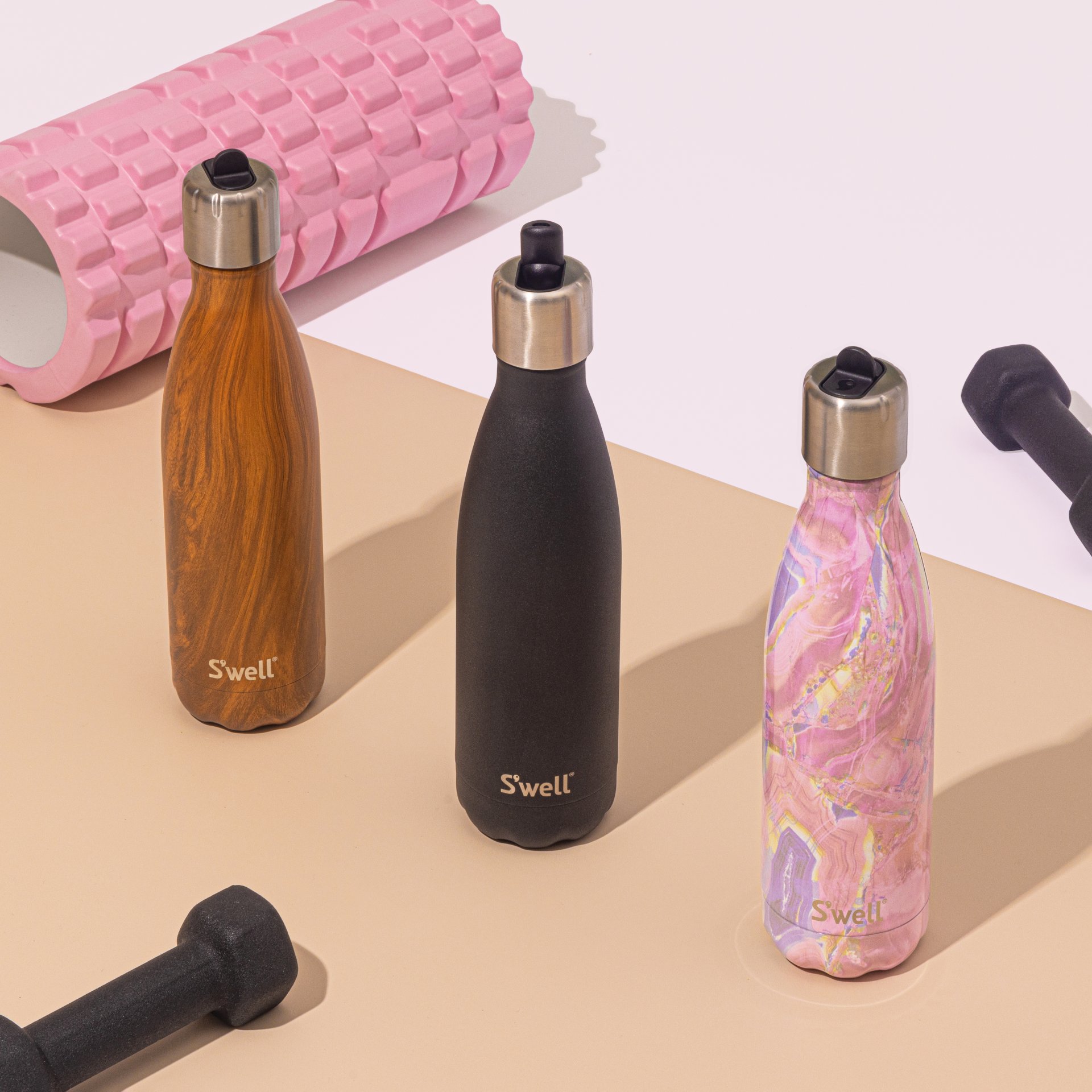 Three different colours of S'Well water bottles are laid out alongside workout equipment.