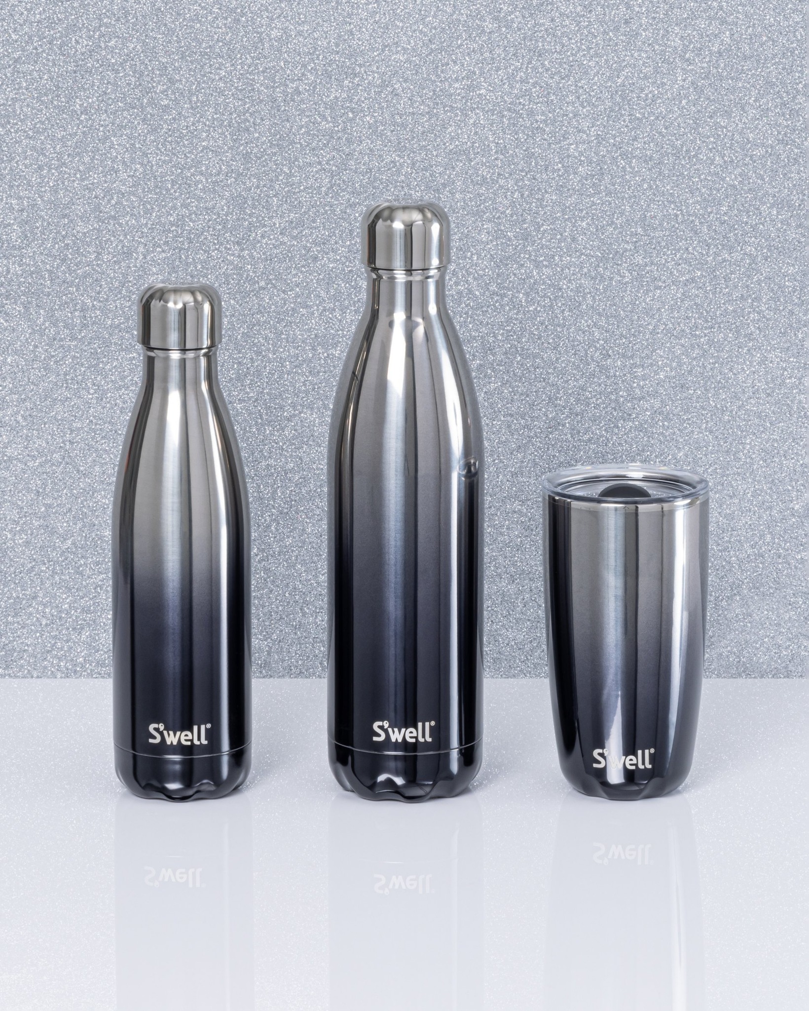 Three different sizes of S'Well water bottles are arranged side by side. They are all the same black to silver ombre design.