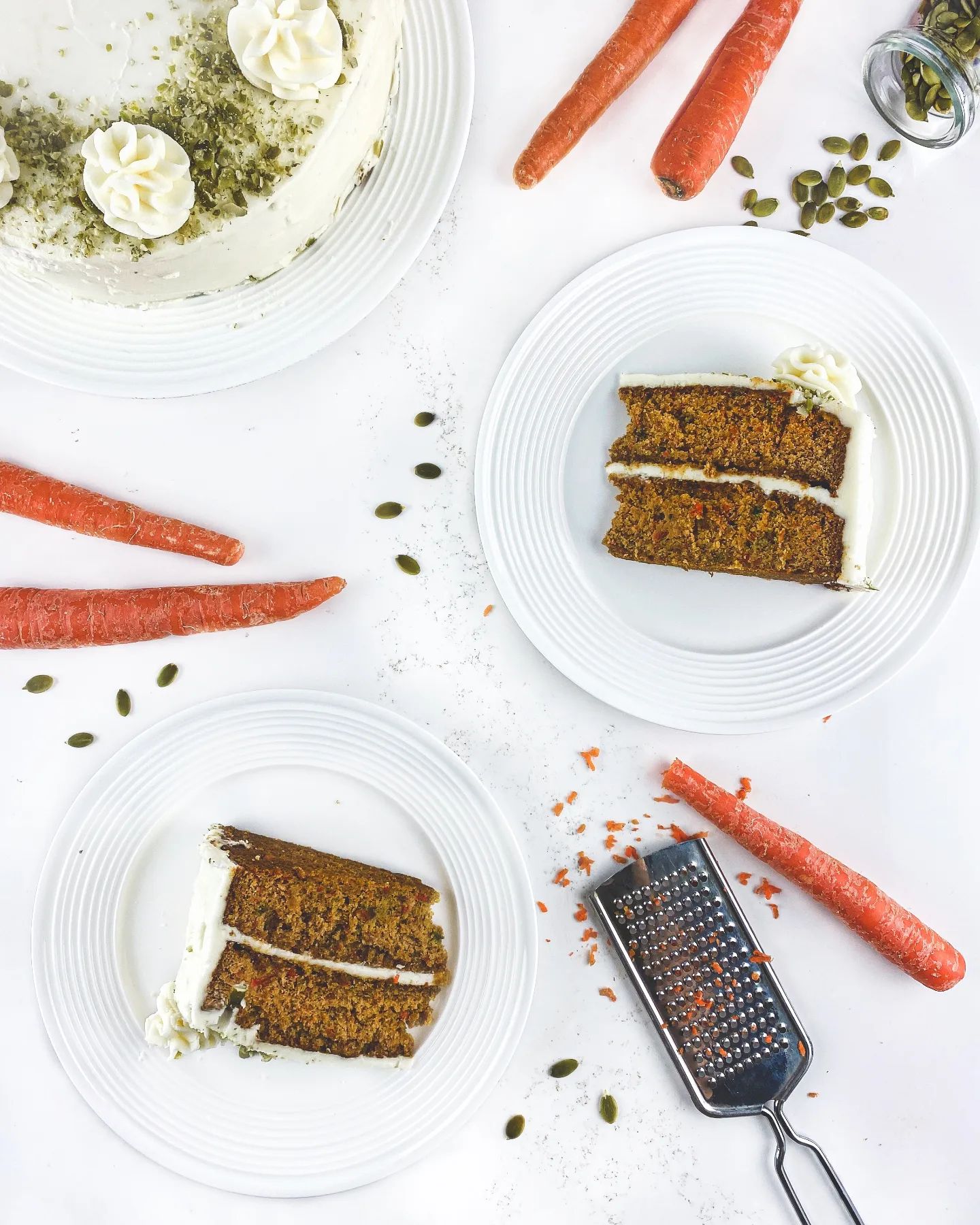 Two slices of carrot cake are displayed on a table alongside shaved carrots and a grater.