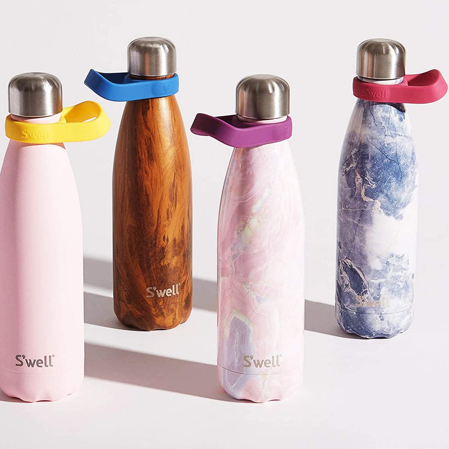 Image of four staggered S'well bottles in various colours: (From left to right): Pink with a silver lid and yellow band around the bottle neck, Wooden-style print with a silver lid and a blue band around the water neck, Pink gemstone-style print with purple, white and gold accents with a silver lid and a purple band around the bottle neck, and blue gemstone with white accents with a silver lid and a red band around the bottle neck.