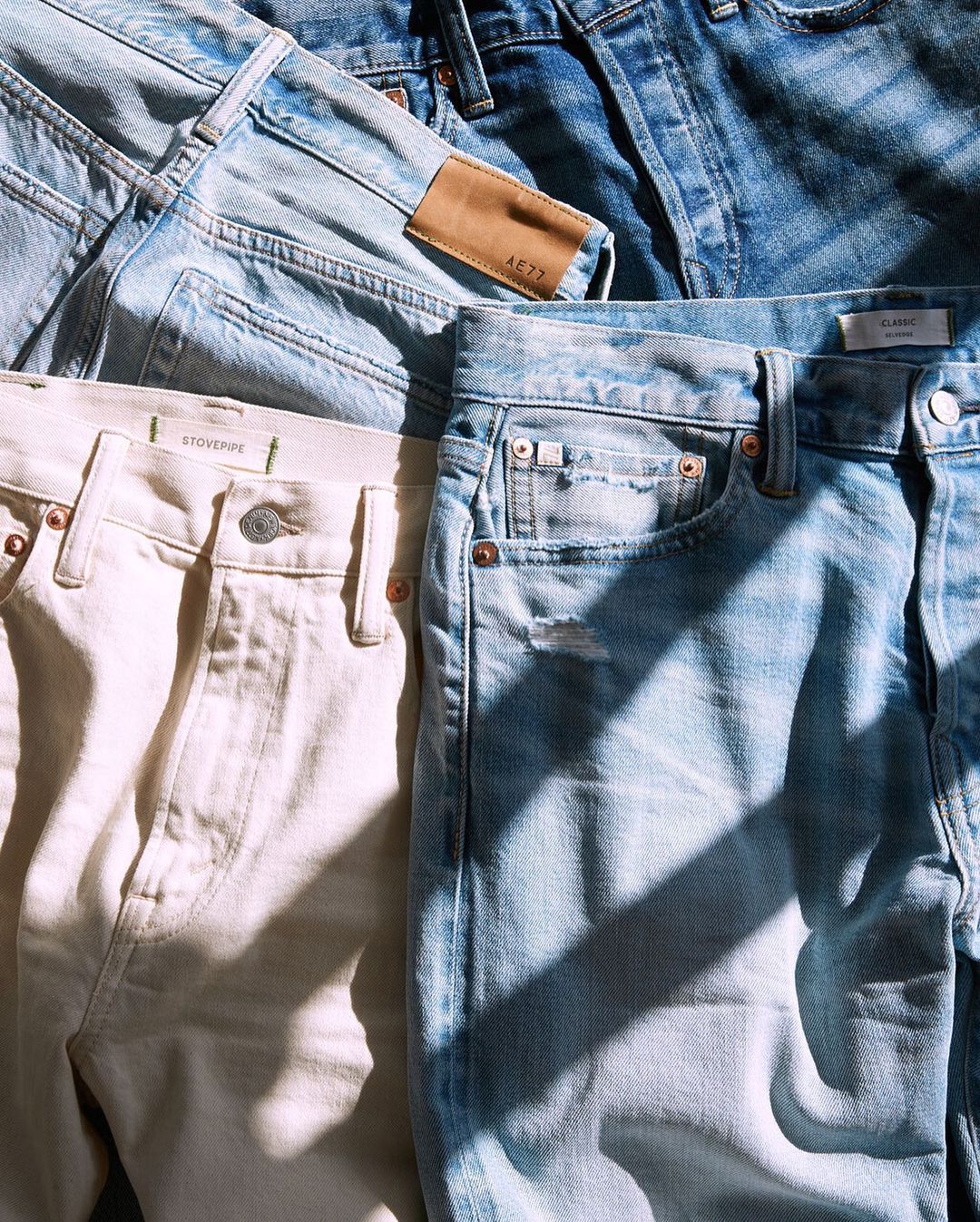 Brightly lit flat lay of jeans stacked on top of each other. On the left are blue and white jeans and on the right are two other blue jeans. A ray of shadows accents the photo.