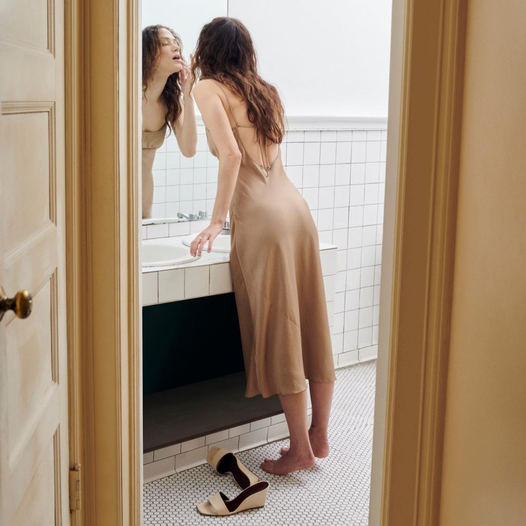 image of a woman looking at herself in a bathroom mirror. she wears a neutral coloured satin dress with a low back