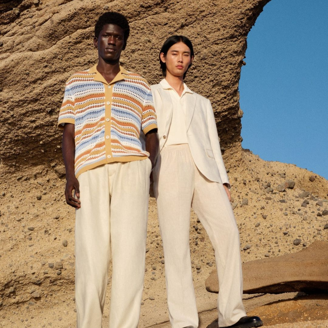 image of two men in a desert. the one on the left wears a striped crochet button up shirt and beige pants. the man on the left wears a light blazer, a white polo shirt and light pants