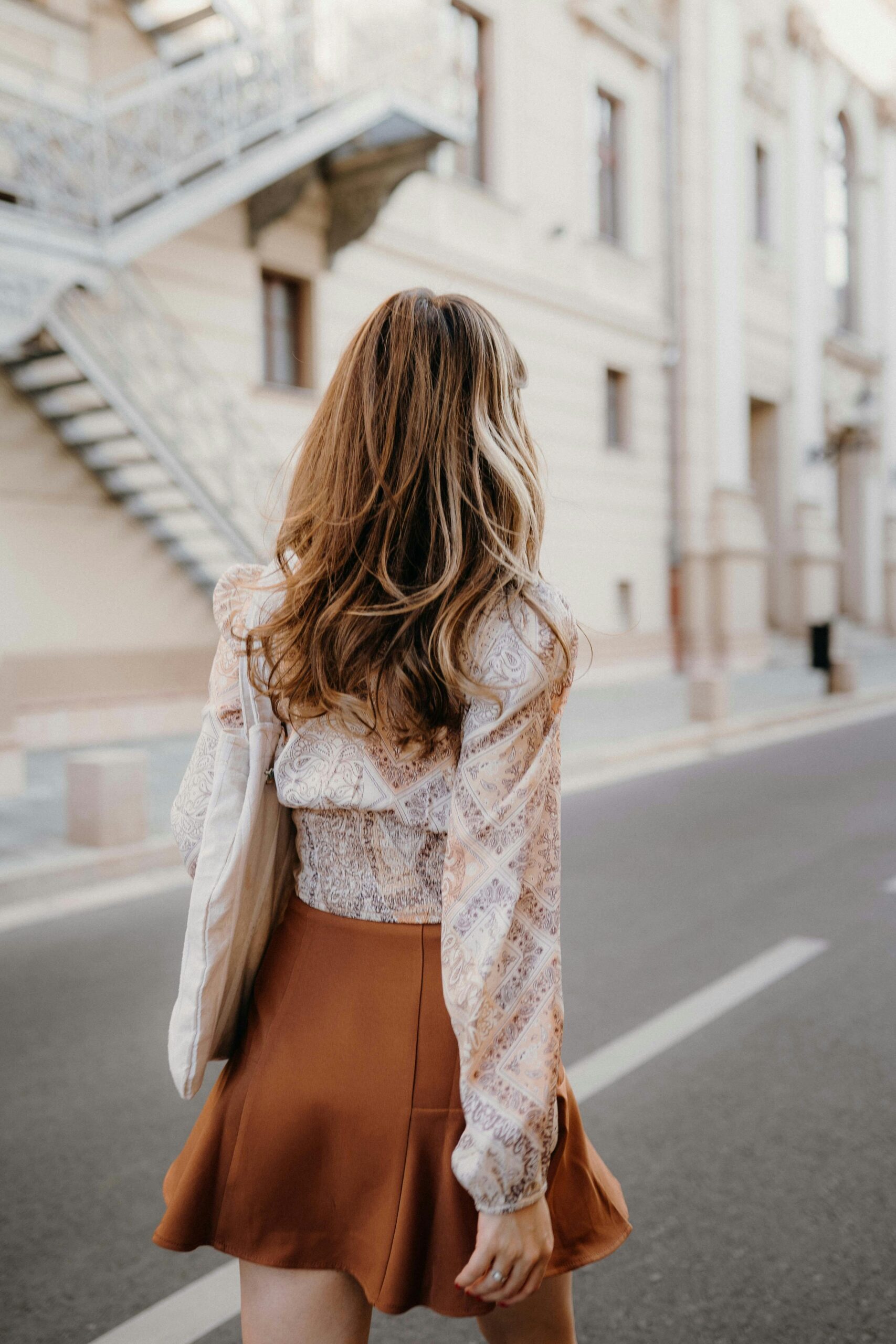 Woman wearing brown skirt and white blouse carrying laptop walking down street.