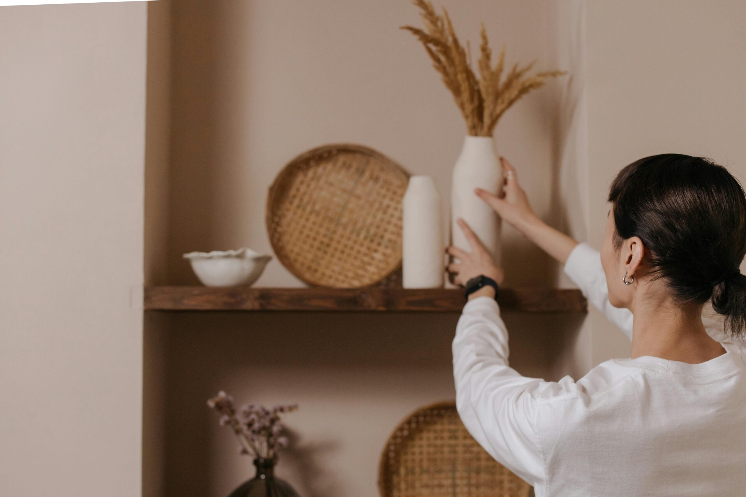 Woman putting white vase on floating brown wood shelf with other decor items.